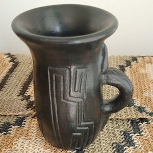 Puede incluir: Una taza de cerámica negra con un borde acampanado y un asa. La taza presenta un diseño geométrico tallado en la superficie. La taza está sobre un textil tejido.