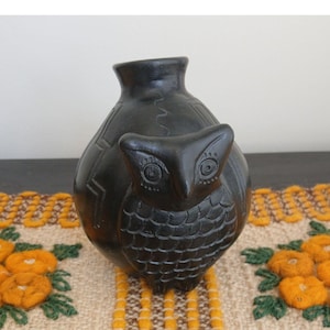 Può includere: Un vaso di ceramica nero a forma di gufo, con una figura di gufo dettagliata sul davanti. Il vaso ha un corpo arrotondato e un collo stretto. Le piume del gufo sono intagliate in modo intricato. Il vaso è appoggiato su un runner decorativo.
