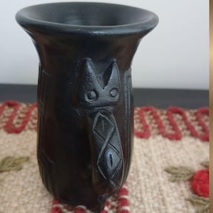 Può includere: Una tazza in ceramica nera con manico e bordo svasato. La tazza presenta motivi geometrici scolpiti e un volto stilizzato di gatto sul manico. La tazza è alta circa 13 cm.