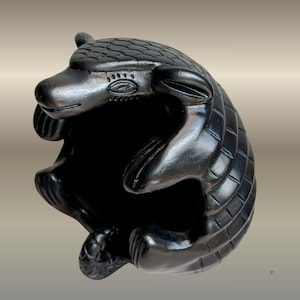 Könnte beinhalten: Schwarze Keramikskulptur eines sitzenden Gürteltiers. Die Skulptur hat eine detaillierte, strukturierte Oberfläche mit einem segmentierten Panzer und einem glatten, abgerundeten Kopf. Das Gürteltier hat kleine Ohren, eine lange Schnauze und einen gebogenen Schwanz.