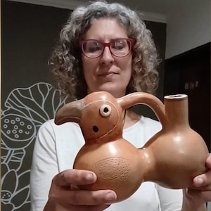 Puede incluir: Un recipiente de cerámica marrón claro con forma de ave, que presenta un pico curvado, un ojo circular y un detalle de ala texturizada. La pieza tiene un cuerpo hueco y redondeado unido a un pico vertical mediante un asa de arcilla mate.