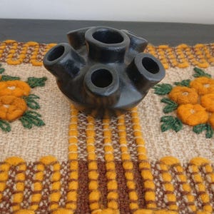 Puede incluir: Un jarrón de cerámica negro con seis aberturas. El jarrón se encuentra sobre un textil tejido con bordados florales naranjas y un patrón de rayas amarillas y marrones.
