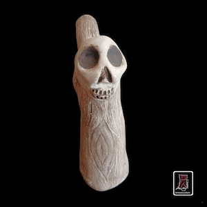 Puede incluir: Una escultura de cerámica de color beige claro con forma de tronco de árbol y una calavera tallada en la parte superior. La calavera presenta cuencas oculares oscuras y huecas y una mandíbula detallada. El tronco tiene líneas verticales.