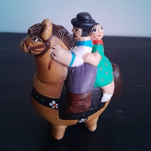 Puede incluir: Figura de cerámica que representa un caballo marrón con dos figuras. El jinete lleva sombrero negro, camisa blanca y pantalón morado. El pasajero lleva un vestido verde con lunares rojos. El caballo tiene una silla de montar negra.