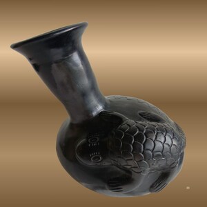 May include: A black ceramic vessel with a long neck and a rounded body. The body of the vessel is decorated with a detailed relief of a turtle. The vessel has a smooth, glossy finish and is tilted slightly to the side.