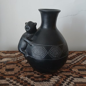 Può includere: Un vaso in ceramica nera con una figura di orso. Il vaso ha un corpo arrotondato e un collo stretto. Motivi geometrici sono scolpiti attorno alla base. L'orso ha dettagli di pelliccia e tratti del viso scolpiti. Circa 20 cm di altezza.