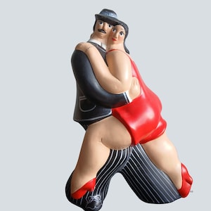 Puede incluir: Una escultura de cerámica de una pareja bailando. El hombre lleva un traje y sombrero negros, mientras que la mujer viste un vestido y zapatos rojos. El pantalón del hombre tiene rayas blancas y negras. La escultura representa un baile de tango.