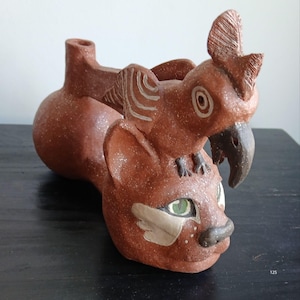 Könnte beinhalten: Handgefertigte Keramikskulptur mit einer katzenartigen Figur mit grünen Augen, gekrönt von einem Vogel. Die rotbraune Keramik hat weiße Sprenkel und steht auf einer dunklen Oberfläche. Der Vogel hat einen dunklen Schnabel und einen strukturierten Kamm.