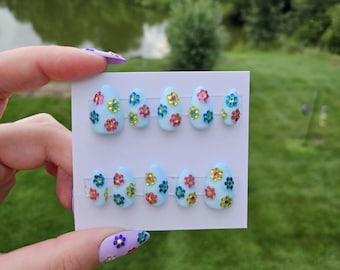 Puede incluir: Uñas postizas de color azul claro con adornos florales coloridos. Cada uña presenta una disposición diferente de flores rosas, amarillas y azules. Las uñas se muestran en una tarjeta blanca, perfectas para manicura.