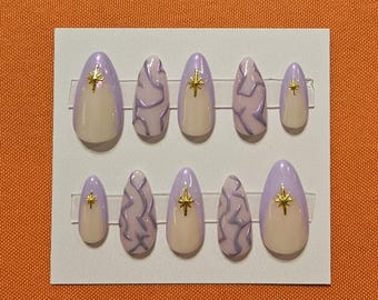 Pode incluir: Um conjunto de dez unhas postiças ovais. Cada unha apresenta uma base lilás clara com uma ponta creme e um detalhe de estrela dourada. Algumas unhas têm um design ondulado lilás claro.