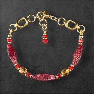 May include: Gold-toned bracelet featuring a mix of red and yellow beads. The bracelet includes elongated red beads with a speckled design, small red beads, and round yellow beads. The clasp and connecting elements are gold-colored.