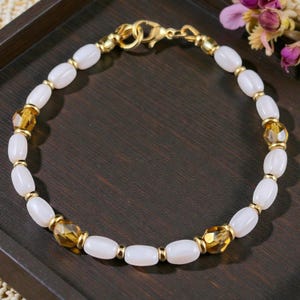 May include: A delicate bracelet featuring oval white beads interspersed with faceted amber-colored beads and small gold-toned accents. The bracelet is secured with a gold clasp and is presented on a dark wooden surface.