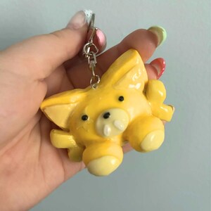 May include: A yellow keychain featuring a stylized animal figure with pointed ears, small black eyes, a snout with two white teeth, and rounded limbs. The figure is attached to a silver-toned metal split ring and a short chain connector.