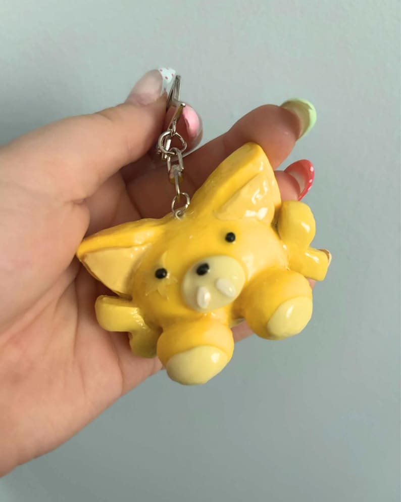 May include: A yellow keychain featuring a stylized animal figure with pointed ears, small black eyes, a snout with two white teeth, and rounded limbs. The figure is attached to a silver-toned metal split ring and a short chain connector.
