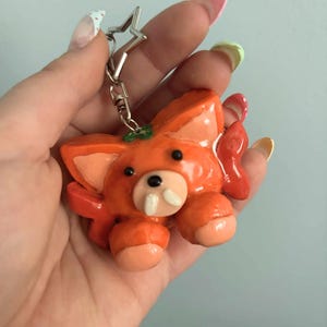May include: A glossy orange fox-like creature keychain with large ears, two small black eyes, and two prominent white fangs. The figure has light peach-colored paws and ears, and it is attached to a silver-colored star-shaped metal keyring clasp.