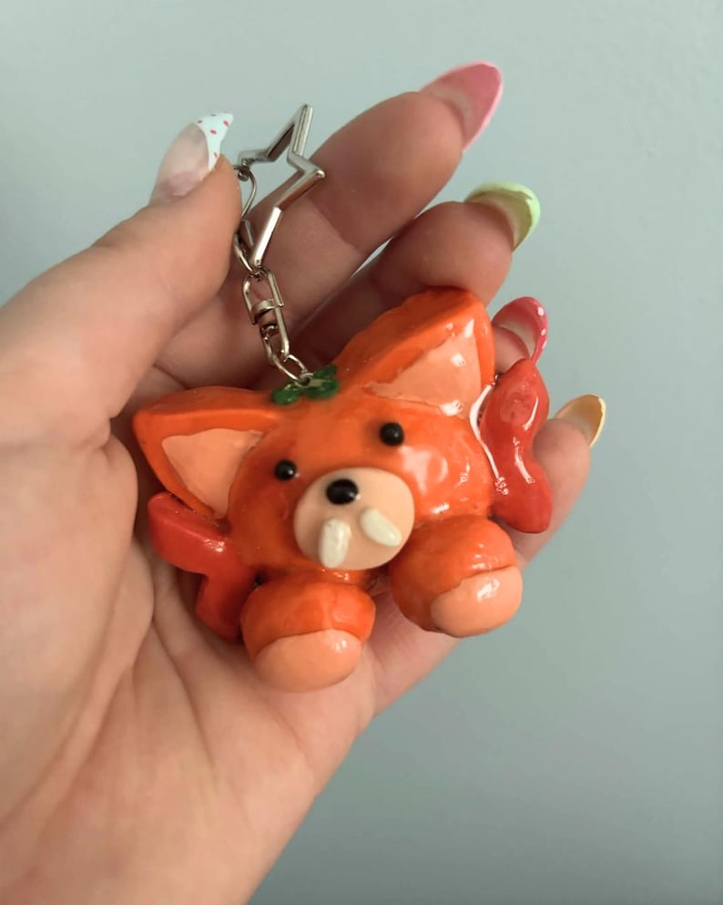 May include: A glossy orange fox-like creature keychain with large ears, two small black eyes, and two prominent white fangs. The figure has light peach-colored paws and ears, and it is attached to a silver-colored star-shaped metal keyring clasp.