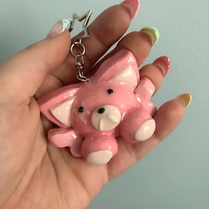 May include: A glossy pink animal-shaped keychain featuring a star-shaped metal ring attachment. The figure has large pointed ears with lighter pink inner details, two small black eyes, and a snout with two white tusks, finished with a smooth coating.