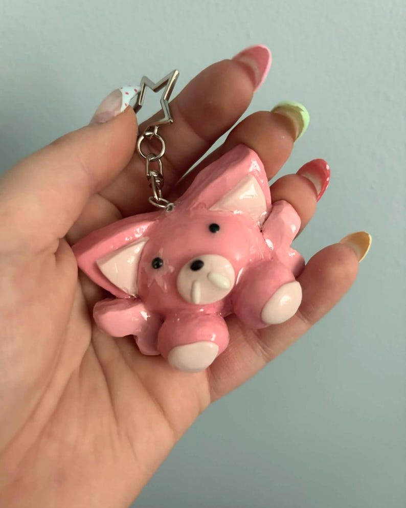 May include: A glossy pink animal-shaped keychain featuring a star-shaped metal ring attachment. The figure has large pointed ears with lighter pink inner details, two small black eyes, and a snout with two white tusks, finished with a smooth coating.