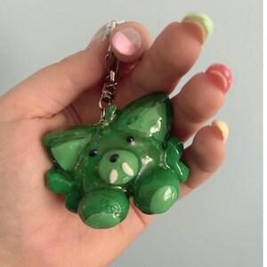 May include: A glossy green keychain shaped like a small creature with pointed ears, a rounded snout, and two small white tusks. The figure has a smooth, shiny finish and is attached to a silver-toned metal lobster clasp and a short chain link.