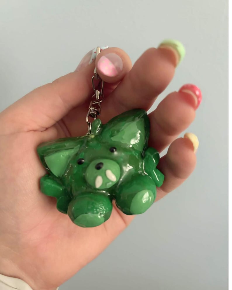May include: A glossy green keychain shaped like a small creature with pointed ears, a rounded snout, and two small white tusks. The figure has a smooth, shiny finish and is attached to a silver-toned metal lobster clasp and a short chain link.