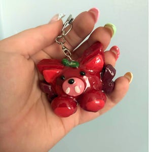 May include: A glossy red keychain featuring a stylized creature with pointed ears, a small black nose, two white tusks, and dark purple wing-like appendages on the sides. The figure has a smooth, shiny finish and is attached to a silver metal keyring.