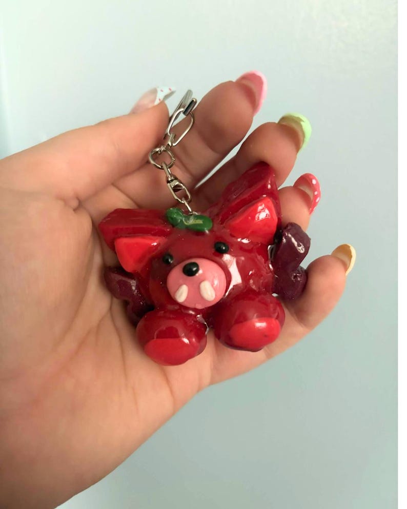 May include: A glossy red keychain featuring a stylized creature with pointed ears, a small black nose, two white tusks, and dark purple wing-like appendages on the sides. The figure has a smooth, shiny finish and is attached to a silver metal keyring.