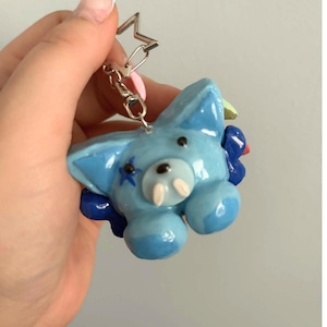 May include: A light blue, glossy, sculpted animal keychain featuring a star-shaped metal ring. The creature has two small white tusks, black dot eyes, a black nose, and a blue star marking on its cheek, accented by dark blue floral elements.
