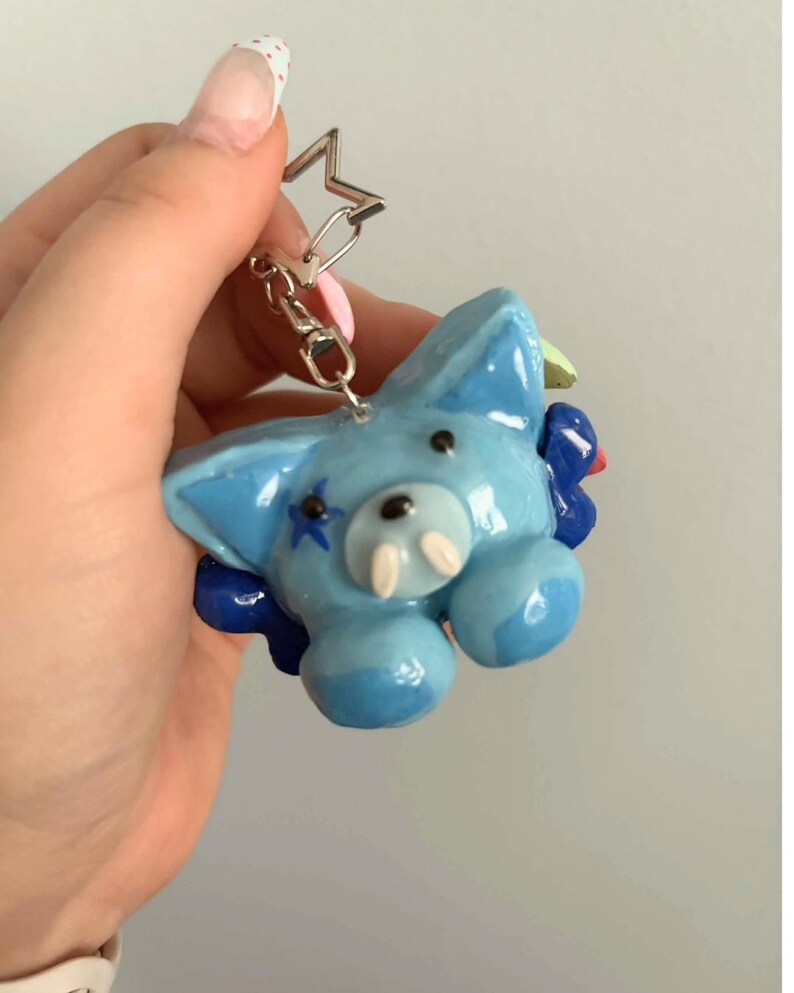 May include: A light blue, glossy, sculpted animal keychain featuring a star-shaped metal ring. The creature has two small white tusks, black dot eyes, a black nose, and a blue star marking on its cheek, accented by dark blue floral elements.