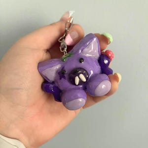May include: A purple creature keychain with a glossy finish, featuring two prominent white fangs, a small black star detail on the side, and a tiny red and white spotted mushroom accent attached to the side of its head with a metal ring.