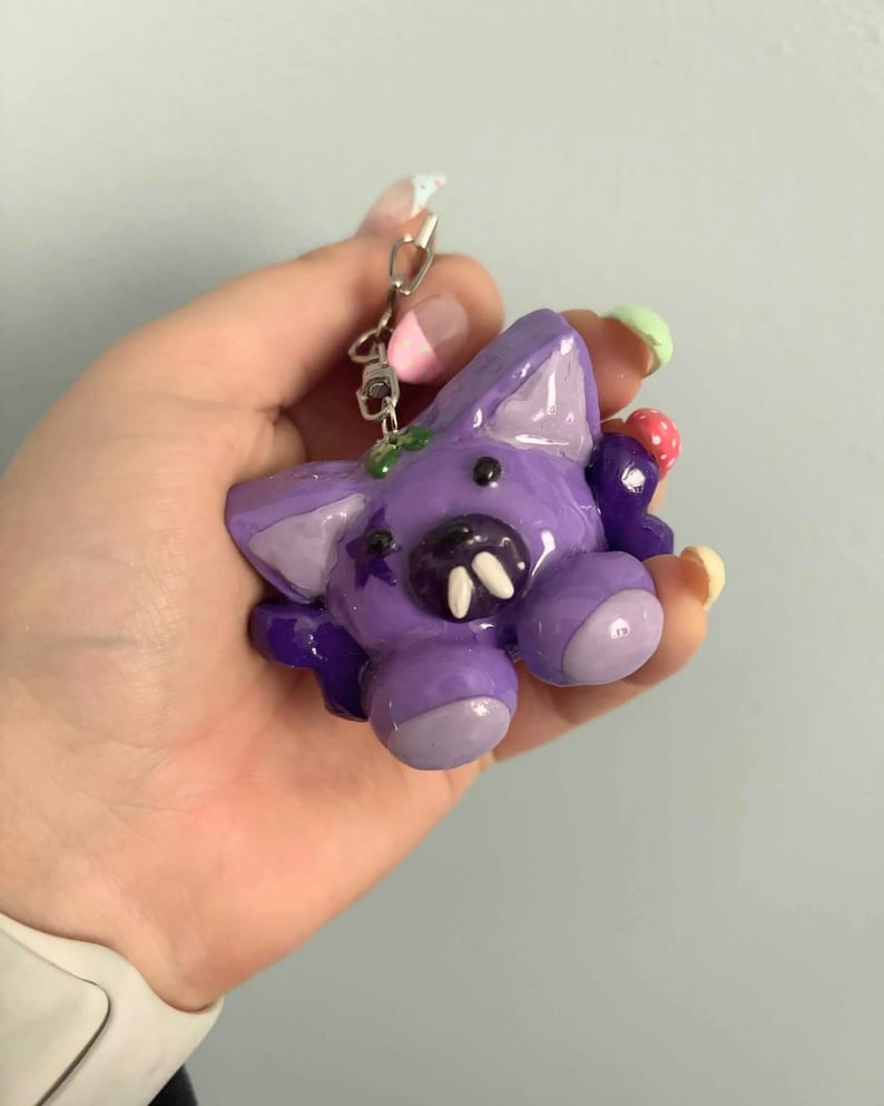 May include: A purple creature keychain with a glossy finish, featuring two prominent white fangs, a small black star detail on the side, and a tiny red and white spotted mushroom accent attached to the side of its head with a metal ring.