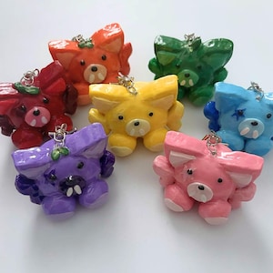 May include: A collection of seven small, glossy, sculpted animal charms in various colors including red, orange, yellow, green, blue, purple, and pink. Each charm features pointed ears, two small white fangs, and a silver metal jump ring for attachment.