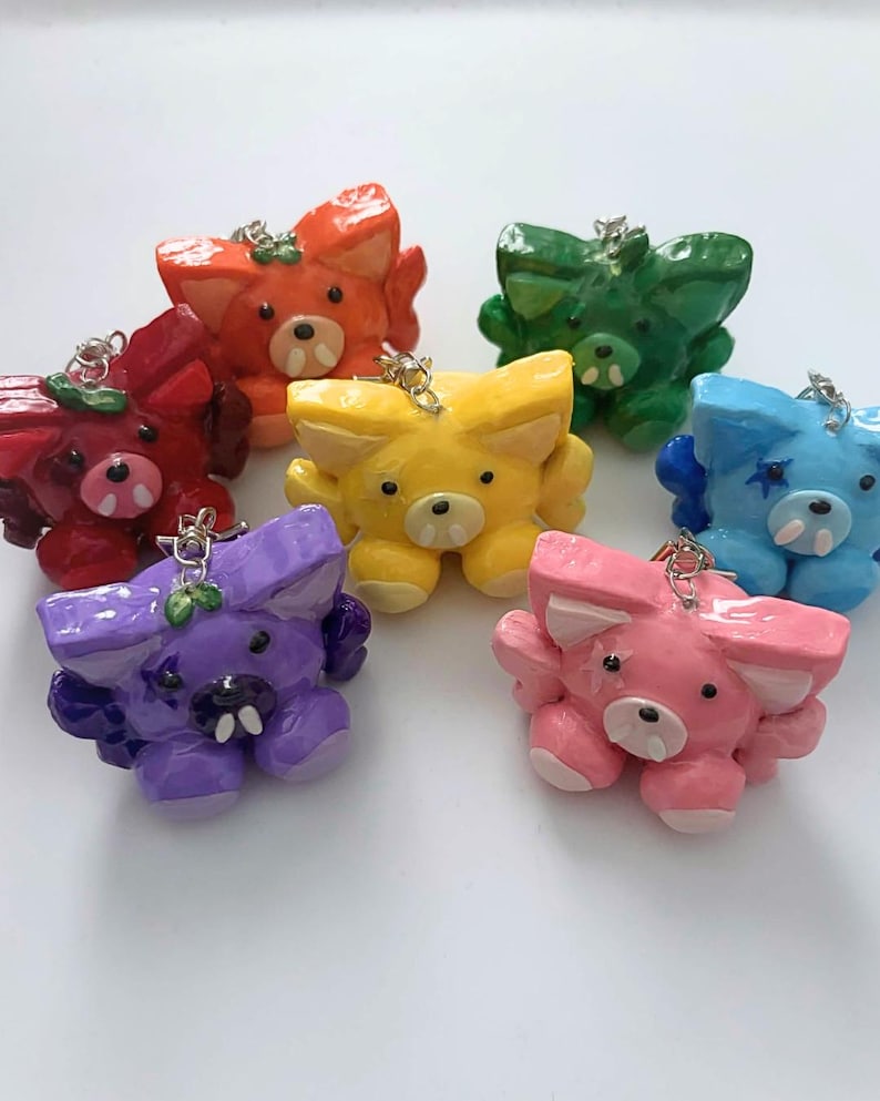 May include: A collection of seven small, glossy, sculpted animal charms in various colors including red, orange, yellow, green, blue, purple, and pink. Each charm features pointed ears, two small white fangs, and a silver metal jump ring for attachment.