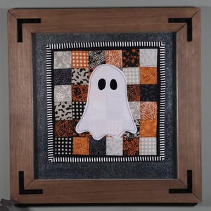 May include: A square wooden frame with black corner accents containing a fabric quilt square. The quilt features a patchwork pattern of orange, black, white, and gray fabrics with spiderweb and pumpkin prints, centered with a white ghost applique.