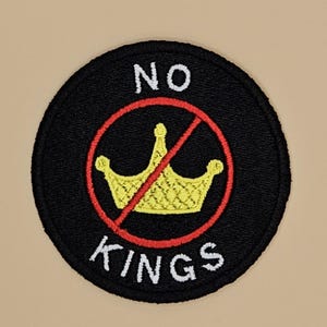 Puede incluir: Parche circular negro con las palabras "NO KINGS" en blanco. Una corona amarilla tachada por una línea roja se encuentra en el centro. El parche es bordado y tiene una apariencia texturizada.
