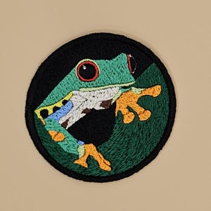 Pode incluir: Patch bordado com uma rã-arborícola verde com olhos vermelhos, patas laranja e barriga branca e azul. A rã está sobre um fundo verde escuro dentro de uma borda circular preta. Ideal para roupas ou acessórios.