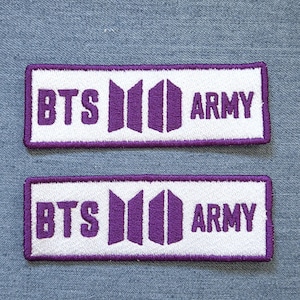 Könnte beinhalten: Zwei rechteckige gestickte Aufnäher mit weißem Hintergrund und violettem Rand. Der Text BTS und ARMY ist in Violett gestickt und durch ein violettes Logo-Symbol getrennt, das aus zwei vertikalen Trapezformen in der Mitte besteht.