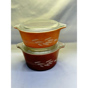 Puede incluir: Dos fuentes de horno Pyrex vintage, una naranja y otra rojo oscuro, apiladas. Cada fuente tiene una tapa de vidrio transparente y un patrón de trigo blanco. La fuente naranja está encima de la fuente roja.