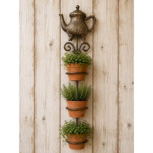 Puede incluir: Una jardinera de pared vertical de metal con una tetera decorativa de tono bronce en la parte superior con adornos de volutas. Debajo de la tetera hay tres anillos metálicos circulares, cada uno sosteniendo una maceta de terracota con plantas verdes.