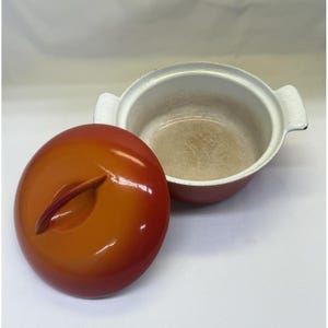 Könnte beinhalten: Ein rot-orangefarbener emaillierter Gusseisentopf mit passendem Deckel. Der Deckel liegt daneben und gibt den Blick auf das weiße Innere des Topfes frei. Der Topf hat weiße Griffe auf beiden Seiten.