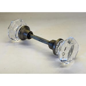 Può includere: Un set di pomelli per porta composto da due pomelli in vetro trasparente, sfaccettati e ottagonali, collegati da un perno metallico centrale. Ogni pomello in vetro è fissato a una rosetta circolare in metallo color bronzo scuro, creando un contrasto tra il vetro trasparente e la finitura metallica scura dei componenti.