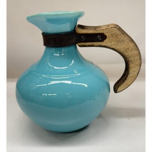 May include: A turquoise ceramic pitcher with a rounded body and a spout. It features a dark brown leather band around the neck and a light-colored wooden handle. The pitcher is likely for serving beverages.