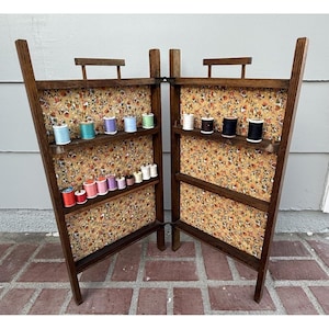 Op de afbeelding: Een vintage houten naaigerei-organizer met meerdere planken, met spoelen gekleurd garen. De binnenkant is gevoerd met bloemenstof. De organizer is open en toont de inhoud tegen een neutrale achtergrond.