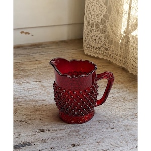 May include: A deep red glass pitcher featuring a textured hobnail pattern across its body. The vessel has a scalloped rim, a curved pouring spout, and a sturdy handle attached to the side. The glass is translucent and catches the light.