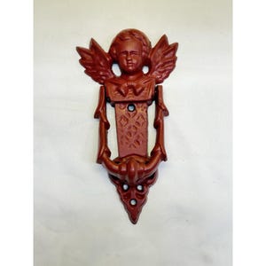 May include: A maroon-colored cast iron door knocker featuring an angel with detailed wings and a decorative bow. The knocker has a detailed design and a classic aesthetic, suitable for exterior doors.