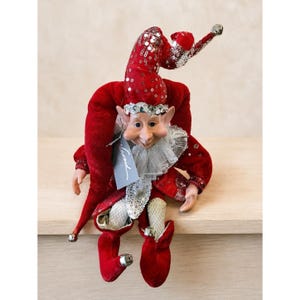 May include: A red velvet jester doll with a smiling face, wearing a sequined hat and collar. The doll has small hands and feet, with silver bells on the hat and shoes. A tag with the word "Carnival" is attached.