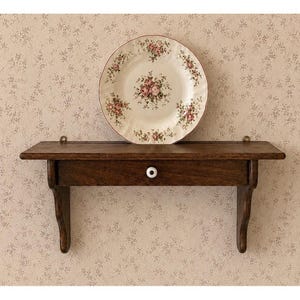 May include: A dark brown wooden wall shelf featuring a single drawer with a small white round knob. Resting on the top surface of the shelf is a round ceramic plate decorated with a pattern of pink roses and green leaves on a cream background.