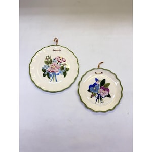 Op de afbeelding: Twee ronde keramische borden met geschulpte randen en groene boorden. Het grotere bord toont een boeket van roze en blauwe bloemen, terwijl het kleinere bord blauwe en paarse viooltjes toont. Beide borden zijn voorzien van metalen ophanghaken.