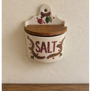 Op de afbeelding: Een aan de muur te bevestigen zoutvaatje van keramiek met een crèmekleurige behuizing en het woord SALT in het rood geschilderd. De houder heeft een houten deksel en een decoratief motief van een rode appel met groene bladeren en bruine takken.