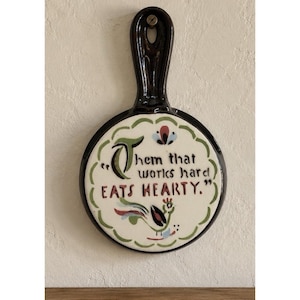 Op de afbeelding: Een ronde, donkerbruine keramische wandplaat met een handvat en een gat om op te hangen. Het midden heeft een crèmekleurig oppervlak met de tekst "Them that works hard EATS HEARTY.", versierd met een kleine, kleurrijke haan en groene rand.