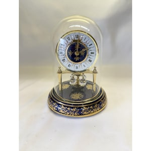 May include: A gold-tone anniversary clock encased in a glass dome, featuring a white dial with Roman numerals, a deep blue center with gold floral accents, and a circular base decorated with intricate gold filigree on a dark blue background.